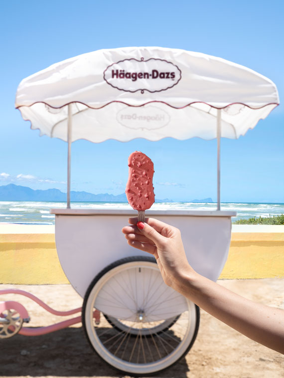 A hand holding a cone of ice cream in front of a Häagen-Dazs ice cream stand on a beach